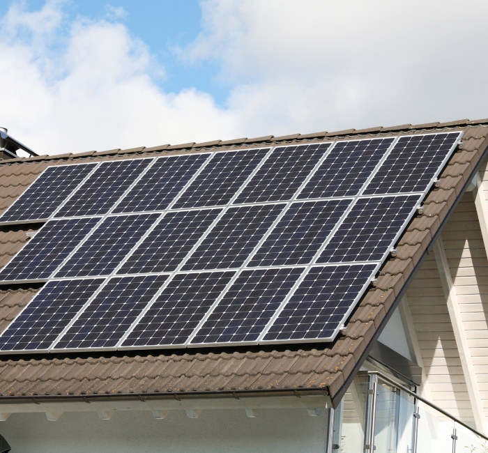 A residential house with a brown tiled roof equipped with solar panels under a partly cloudy sky.